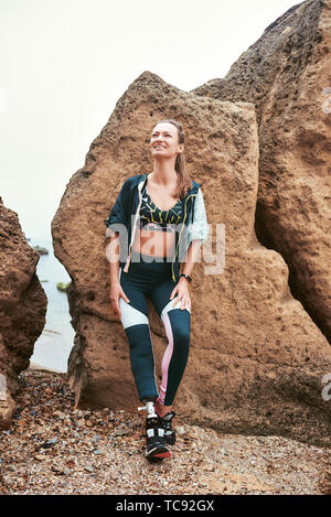 Simplement heureux. Motivé et positif femme handicapée dans les vêtements de sport avec prothèse appuyé contre une pierre sur la plage . Mobilité sportif. Mode de vie sain. Concept de bonheur Banque D'Images