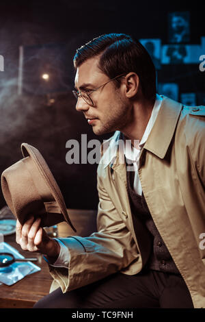 Détective pensive, dans les verres et trench coat assis sur table et holding hat Banque D'Images