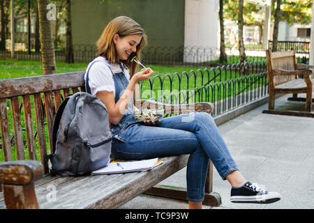 Jeune fille étudiante en salopette denim avoir dîner sain tout en étant assis sur un banc à l'extérieur Banque D'Images