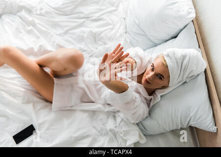 Top View of woman in bijoux avec des serviettes sur la tête et mains tendues couché dans un lit blanc smartphone près de Banque D'Images