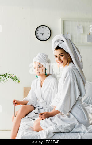 Femmes élégant souriant dans des peignoirs et des bijoux, avec des serviettes sur la tête assis sur le lit avec les limes à ongles et looking at camera Banque D'Images