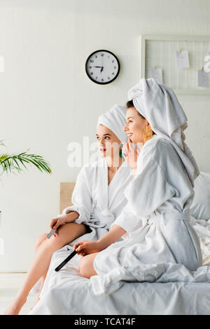 Femmes élégant souriant dans des peignoirs et des bijoux, avec des serviettes sur la tête assis sur le lit avec les limes à ongles et à l'écart Banque D'Images