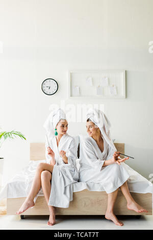 Smiling women élégant dans des peignoirs et des bijoux, avec des serviettes sur la tête assis sur le lit avec les limes à ongles Banque D'Images