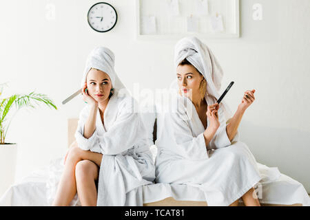 Les femmes élégantes dans des peignoirs et des bijoux, avec des serviettes sur la tête assis sur le lit avec les limes à ongles Banque D'Images