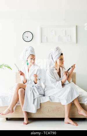 Les femmes aux pieds nus dans des peignoirs et des bijoux, avec des serviettes sur la tête à l'aide de limes à ongles alors que sitting on bed Banque D'Images