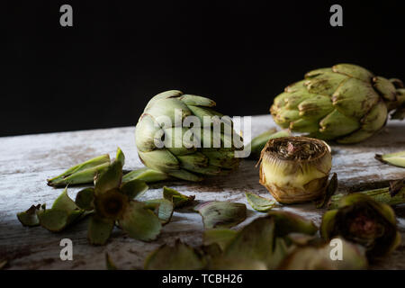 Libre de certains artichauts frais cru entier et certains autres couper les artichauts et leurs feuilles sur une table rustique en bois blanc, sur un fond noir Banque D'Images