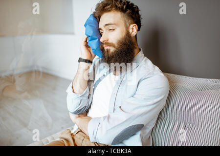 Homme barbu avec des maux de l'application de glace tout en étant assis sur le sac à la maison de la table Banque D'Images