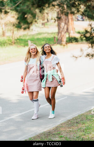 Sur toute la longueur de deux amis multiculturelle avec penny boards embrassant tout en marchant sur la route Banque D'Images