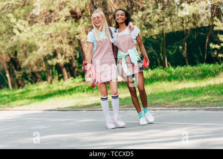 Sur toute la longueur de deux amis multiculturelle avec penny boards embrassant tout en marchant sur la route Banque D'Images