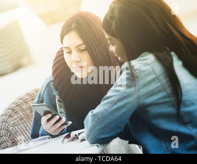 Deux jeunes belles filles s'asseoir à une table et parler en utilisant le téléphone mobile. Banque D'Images