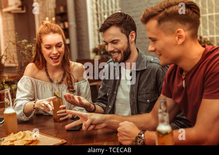 Des boissons avec des amis. Rousse charmante jeune femme séduisante et deux gars attrayant rayonnant heureux assis au bar avec boissons et s'amusant Banque D'Images