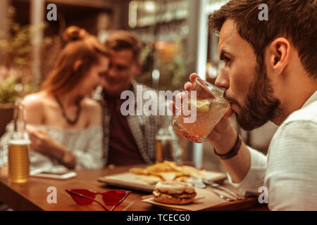 Se sentir comme la troisième roue. Close-up face portrait de barbu brun beau jeune-homme adulte seul sirotant un cocktail pendant que ses camarades hugging Banque D'Images