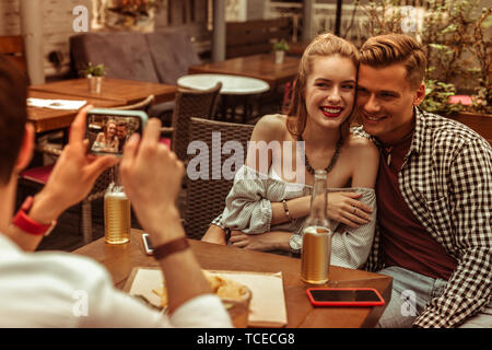 Heureux couple. Heureux l'amour belle belle-appelant les jeunes-adultes smiling hugging couple rayonnant photographié par leur ami au bar. Banque D'Images