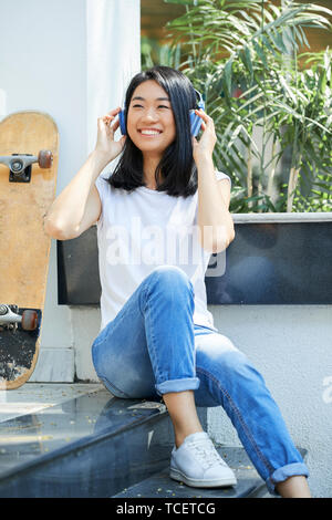 Vietnamiens positive teenage girl sitting on steps et écouter de la musique dans ses écouteurs Banque D'Images