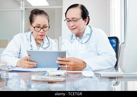 Les travailleurs médicaux asiatiques expérimentés en laboratoire assis à table et discuter de l'histoire médicale du patient sur tablet computer Banque D'Images