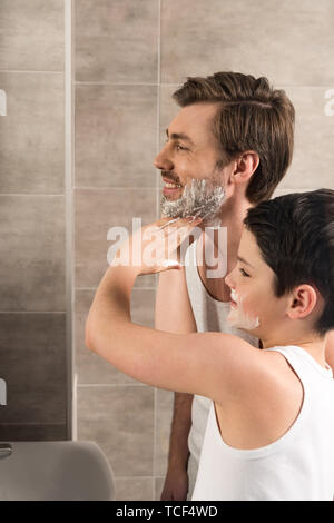Preteen boy l'application de mousse à raser à sourire père dans la salle de bains Banque D'Images