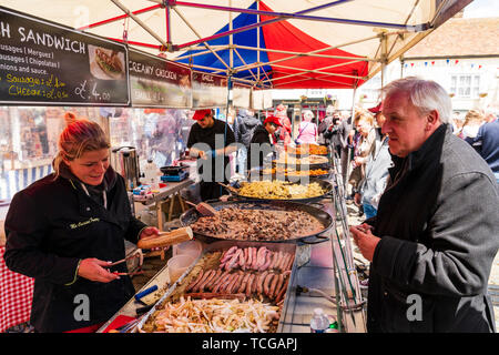 L'événement le week-end à la ville médiévale de Sandwich, en Angleterre. Marché français sont tenus annuellement dans la place de la ville. Vue le long long hot food vente de paella, saucisses français et divers autres aliments cuits dans de grandes casseroles sur le comptoir. Femme Homme senior avec baguette roll avec saucisses dans. Banque D'Images