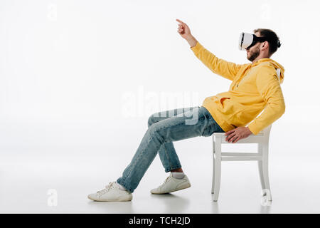 Vue latérale du casque de réalité virtuelle à l'homme assis sur une chaise et en pointant avec le doigt sur fond blanc Banque D'Images