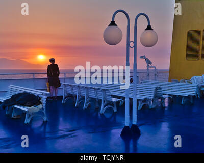 Jeune femme sur le navire à la recherche au coucher du soleil, pont bleu jaune blanc bancs, cheminée, île lointaine montagnes, Mer Égée, Grèce. Banque D'Images