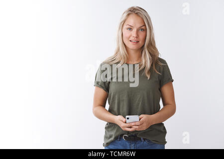 Studio shot of attractive et agréable femme journaliste blonde conversation d'enregistrement via smartphone holding mobile phone près de la poitrine et le regard sur cam Banque D'Images