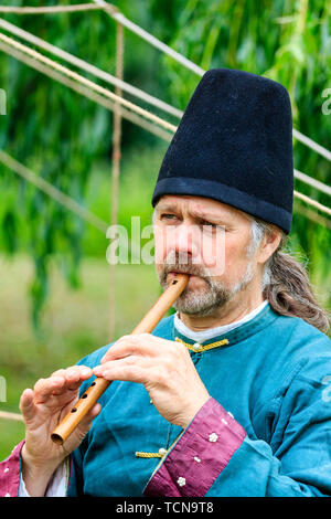 La reconstitution médiévale événement. Portrait d'homme barbu en tunique bleu et noir hat jouer une mélodie sur une petite flûte, à l'extérieur. Tête et épaules. Banque D'Images