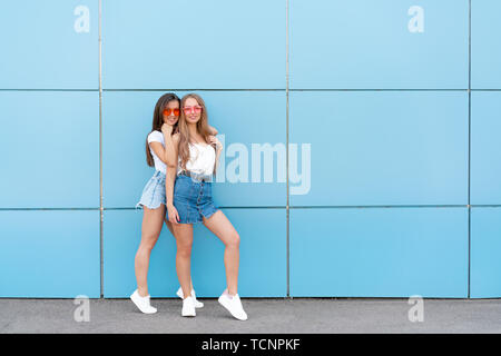 Deux jeunes amis femme hipster en rétro lunettes néon et permanent smiling over blue wall Banque D'Images