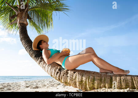 Young Woman in Bikini Sitting On Palm Tree Trunk de boire l'eau de noix de coco At Beach Banque D'Images