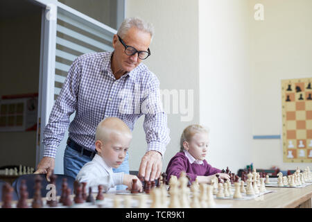 Enseignant de sexe masculin âgés avec des lunettes aide deux élèves avec l'apprentissage des combinaisons d'échecs au cours de class Banque D'Images