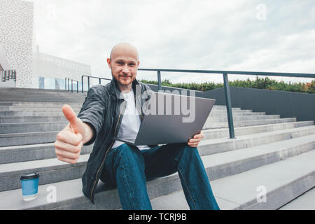 Sourire succès adultes homme barbu chauve en veste noire à l'aide d'ordinateur portable dans des escaliers à ville, montrer Thumbs up Banque D'Images