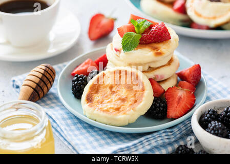 Crêpes au fromage cottage frit syrniki servi avec des fraises, des mûres, du miel et une tasse de café. Serviette à carreaux fond bleu Banque D'Images