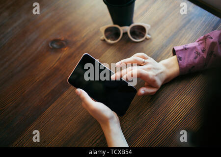 Sommaire des mains de femme, défilement de l'écran du téléphone mobile. Une tasse de café et verres à côté. Banque D'Images