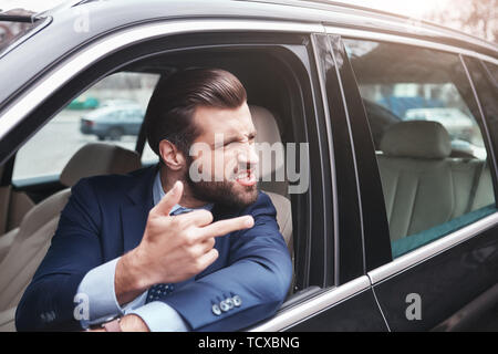 Homme grossier. En colère et rude businessman barbu en tenue de cérémonie est un geste et faire une grimace alors qu'il conduisait sa voiture. Les émotions négatives. Affaires Banque D'Images