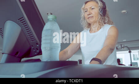 Senior woman est engagé sur un tapis roulant dans la salle de sport. blonde écoute de la musique avec des écouteurs sur le tapis roulant Banque D'Images