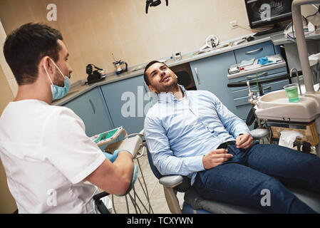 Dark-haired barbu portant des hygiénistes dentaires uniforme blanc est prêt à contrôler les dents du patient. Il est assis près de fauteuil dentaire parle à son patient. La COT Banque D'Images