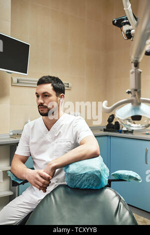 Dark-haired barbu portant des hygiénistes dentaires uniforme blanc est prêt à contrôler les dents du patient. Il est assis près de fauteuil dentaire lors de l'attente pour le patient Banque D'Images