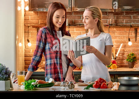 Portrait de deux femmes attrayants, s'amuser, tout en préparant la salade. Ils sont pleinement impliqués dans le processus. Fille blonde en T-shirt blanc et son obscurité Banque D'Images