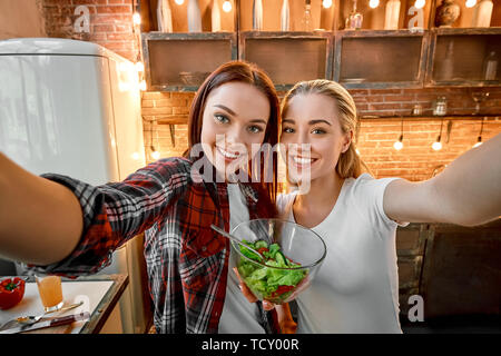 Deux femmes de selfies joyeux, avoir du plaisir, le temps de préparer une salade de légumes. Dark-haired girl wearing chemise à carreaux est titulaire d'une salade cuite pendant que son blo Banque D'Images