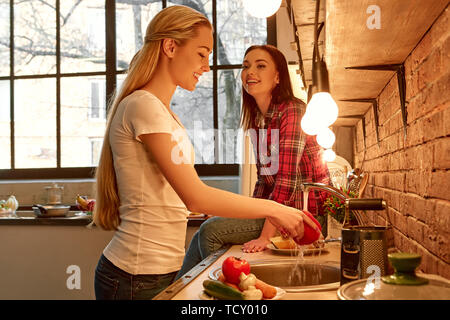 Portrait de deux femmes, s'amuser, tout en préparant une salade de légumes. Les femmes sont en train de rire, de parler les uns aux autres. Blonde girl in white T-shirt se lave veg Banque D'Images