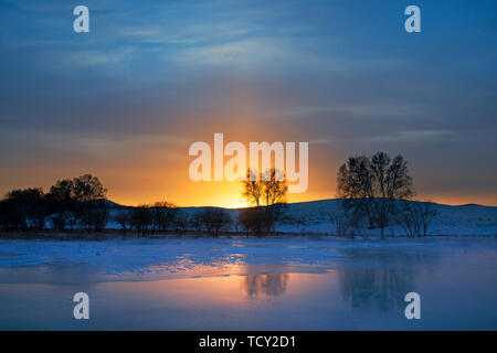 La rivière le lever du soleil. Sur le bord de la rivière sur le barrage de la Mongolie intérieure, tôt le matin de l'hiver, le soleil est encore sous l'horizon, le soleil doré brille dans le ciel, la lueur se reflète sur la glace de la rivière, et le reflet de la petit arbre est également associé à la glace, formant un beau paysage de l'hiver le lever du soleil. Banque D'Images