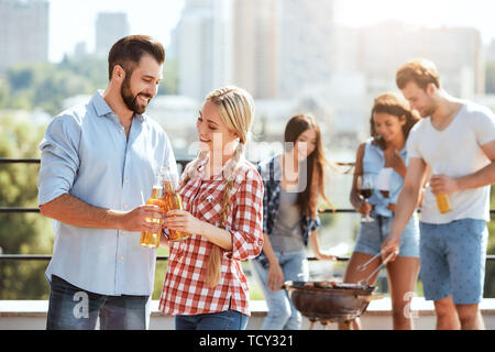 La célébration de l'été. Deux jeunes amis heureux clinking glasses avec bière et souriant alors qu'il se trouvait sur le toit avec des amis. Concept de barbecue. La cuisson. Banque D'Images