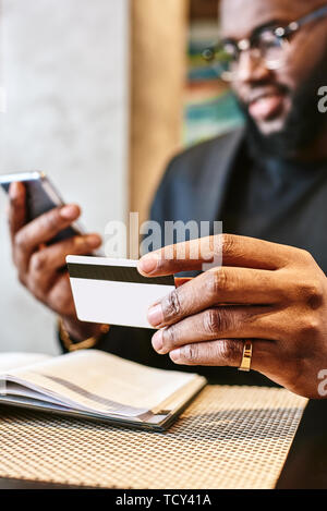 Cropped shot of African-American male holding cell phone dans une main et une carte de crédit dans d'autres, faisant de transaction, à l'aide d'application mobile banking au cours de lun Banque D'Images