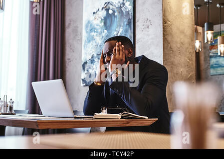 Directeur du bureau d'afro-américaine centrée en bleu foncé veste assis à table de café avec un ordinateur portable, la lecture des documents importants avec expression perplexe et Banque D'Images