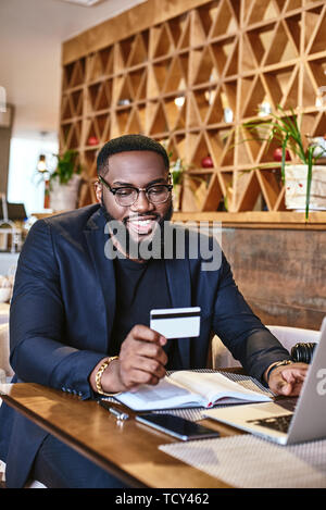 Cropped shot of African-American male holding credit card, faisant de transaction. Ordinateur portable et ses documents de travail sont sur la table. Boutique en ligne et paiement Banque D'Images