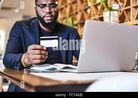 Cropped shot of African-American male holding credit card, faisant de transaction. Ordinateur portable et ses documents de travail sont sur la table. Boutique en ligne et paiement Banque D'Images