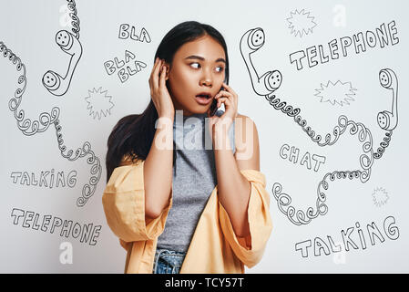 Oh mon Dieu ! Portrait de femme asiatique surpris dans des vêtements décontractés en conversation sur son téléphone intelligent en étant debout contre l'arrière-plan gris avec différents Banque D'Images