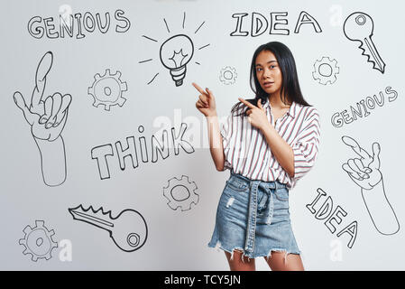 J'ai beaucoup d'idée ! Cute asian woman with long hair pointing at lightbulb debout contre l'arrière-plan gris avec des esquisses à la main sur elle. Idée co Banque D'Images