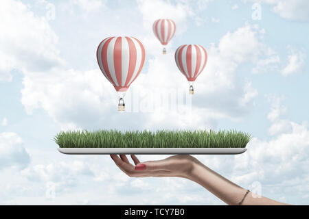 Vue latérale du woman's hand holding digital tablet avec l'herbe verte à l'écran contre le ciel avec des nuages blancs et trois ballons à air chaud à rayures sur elle. Banque D'Images