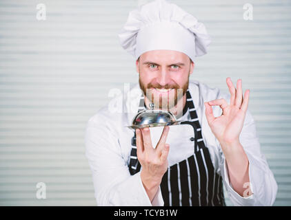 Le cuisinier en chef gesticulant ok signe. Master chef au service de repas au restaurant. Bel homme en tablier et chapeau de cuisinier. Chef cuisinier en uniforme debout avec plat délicieux. Notre restaurant pour tous les goûts. Banque D'Images