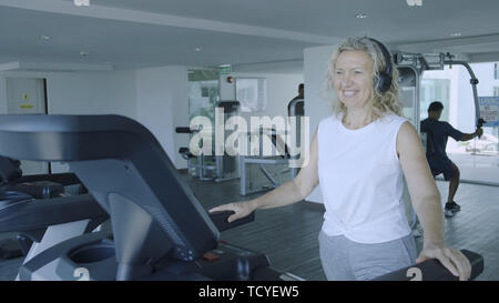 Senior woman est engagé sur un tapis roulant dans la salle de sport. blonde écoute de la musique avec des écouteurs sur le tapis roulant. Banque D'Images