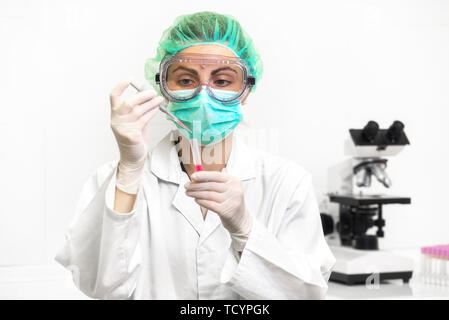 Young attractive concentrée female scientist in lunettes de protection, gants et masque l'abandon d'une substance liquide rouge dans le tube à essai avec une pipette dans le laboratoire . Banque D'Images